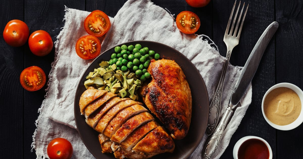 A plate of sliced roasted chicken breast with peas and diced avocado, seen from above, beside small bowls of sauce and halved tomatoes.