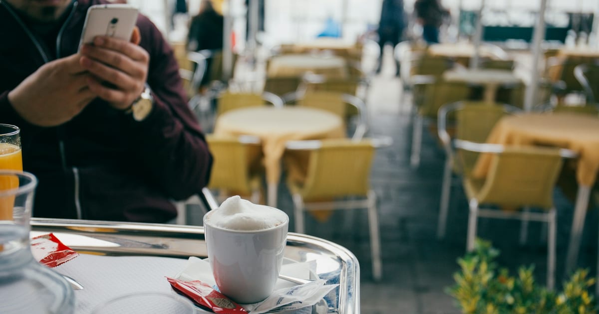 A person holds a smartphone at a cafe table, with a cappuccino and a glass of orange juice in front of them.