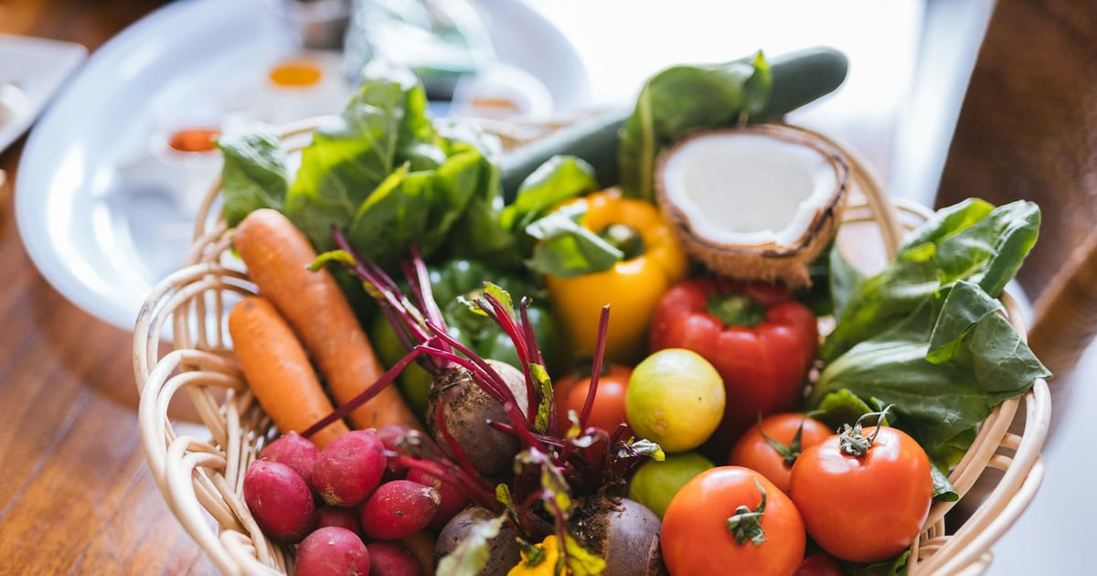 A wicker basket of fresh carrots, radishes, beets, tomatoes, peppers, leafy greens and half a coconut on a wooden table.