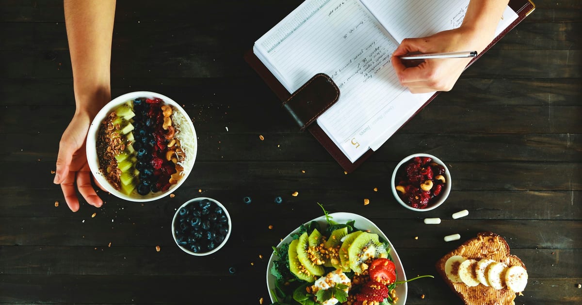 Overhead view of a dark wooden table with a smoothie bowl, a fruit salad and toast with banana, while one hand writes in an open notebook.