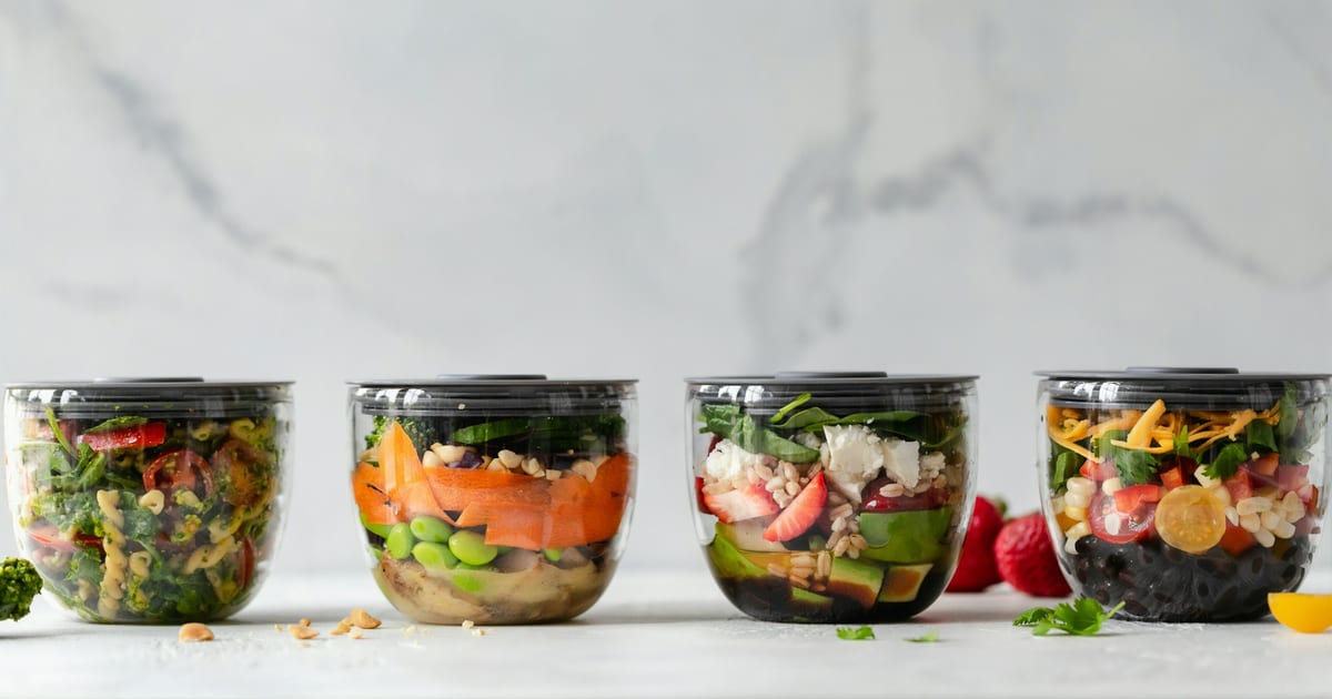 Four clear meal prep bowls in a row on a white counter, filled with pasta salad, edamame and carrots, berries with feta and greens, and black beans with corn and cheese.