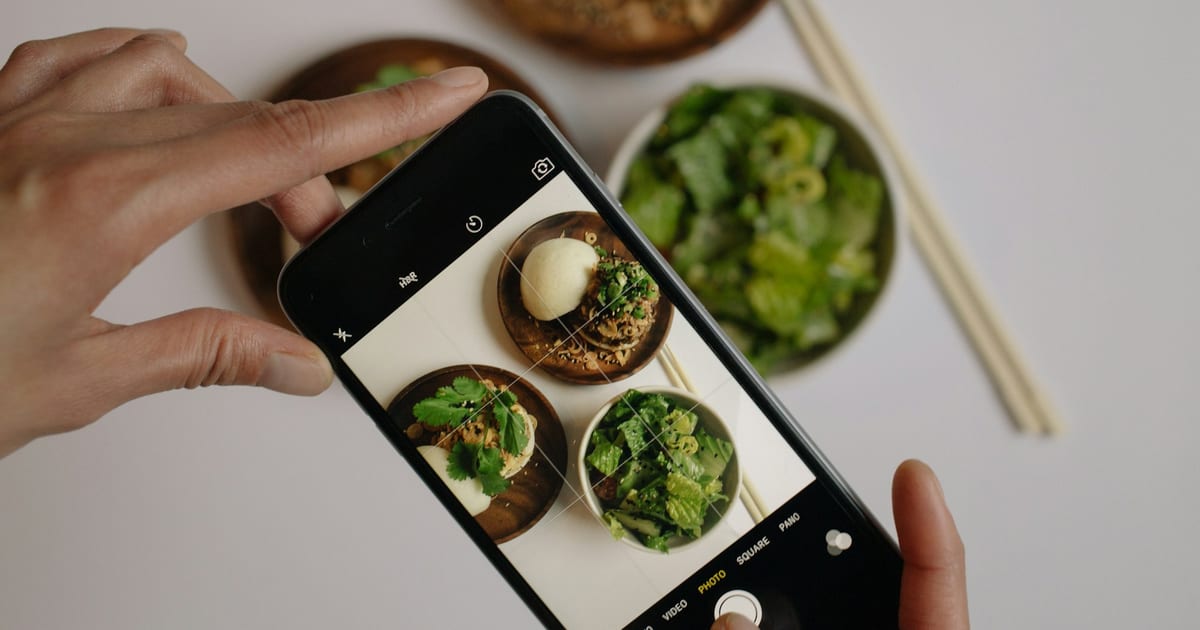 Hands hold a smartphone over a table, framing a photo of two small wooden plates of food and a bowl of green salad.