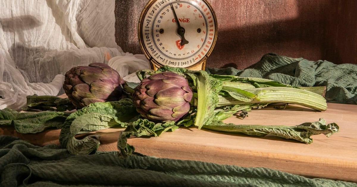 An old dial kitchen scale stands behind two purple artichokes and a bunch of chard on a wooden cutting board.