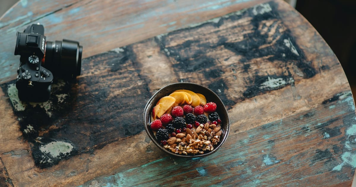 A dark bowl of peach slices, raspberries, blackberries and chopped nuts on a weathered wooden table, next to a camera.