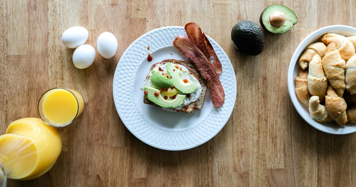 A plate of toast topped with a fried egg and avocado slices, with bacon, next to whole eggs, an avocado, orange juice and a bowl of crescent rolls on a wooden table.