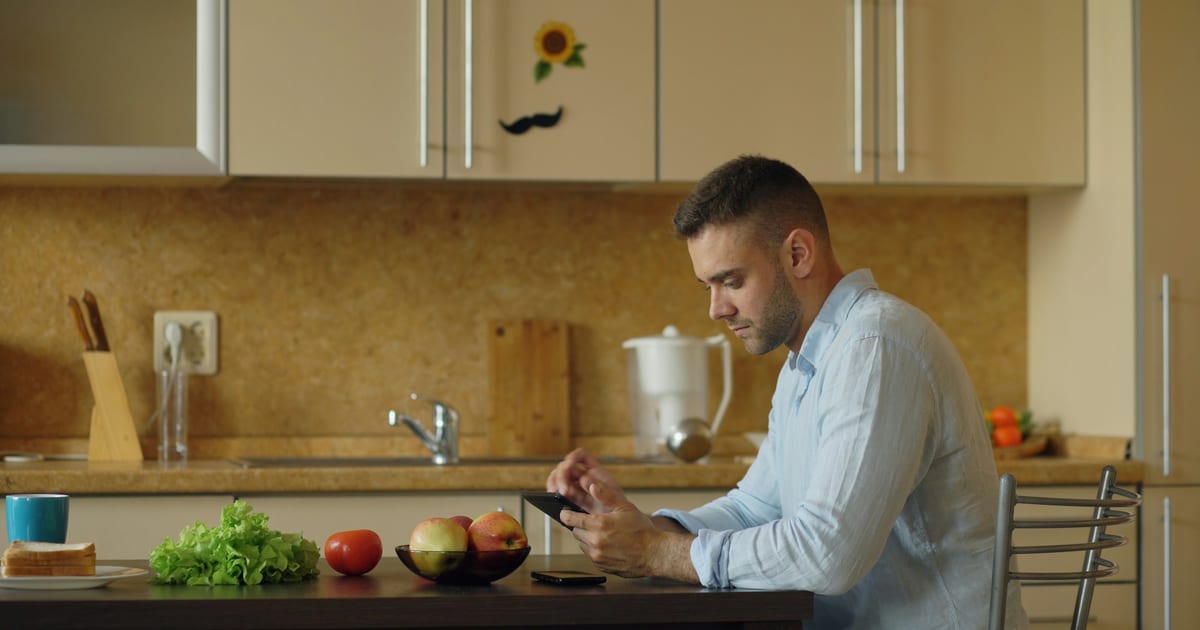 A man sits at a kitchen table with a phone in his hands, next to a bowl of apples, a tomato and a head of lettuce.