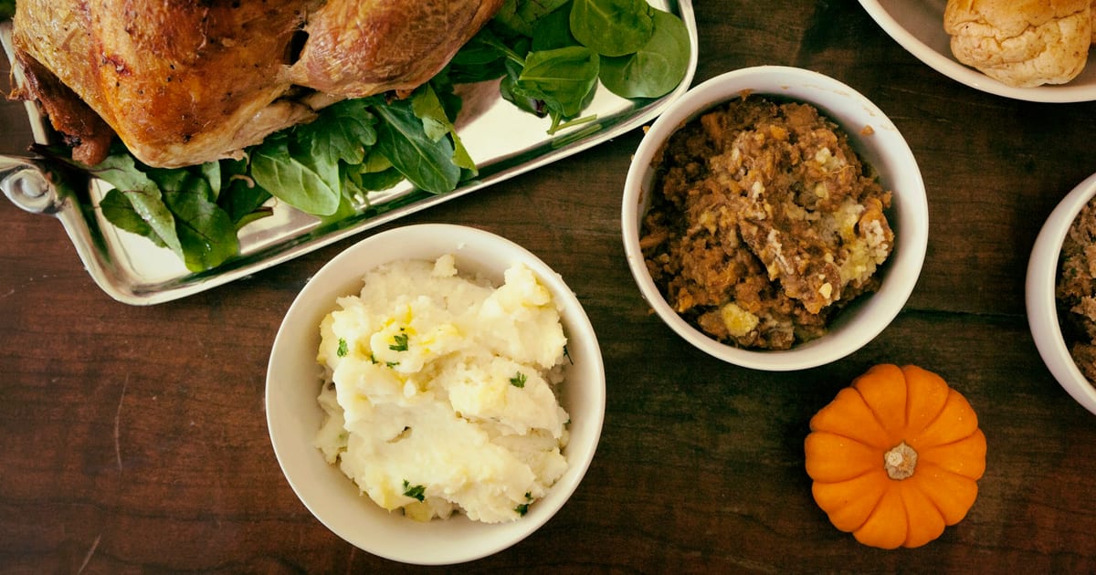 A roast turkey on a silver tray with spinach, beside bowls of mashed potatoes and stuffing, a small pumpkin and a dinner roll on a wooden table.